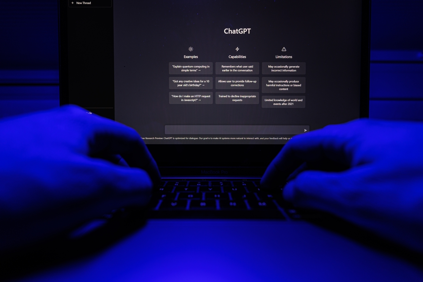 A photo of hands in a dimly lit room with a blue glow typing on a laptop,the screen displays ChatGPT