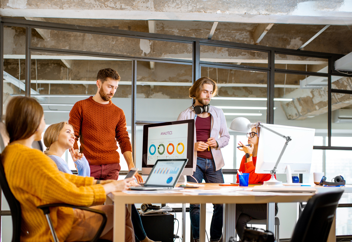 A group of young professionals in a modern office discussing analytics displayed on a computer screen. The atmosphere is collaborative and focused, with charts and graphs visible on the screen.