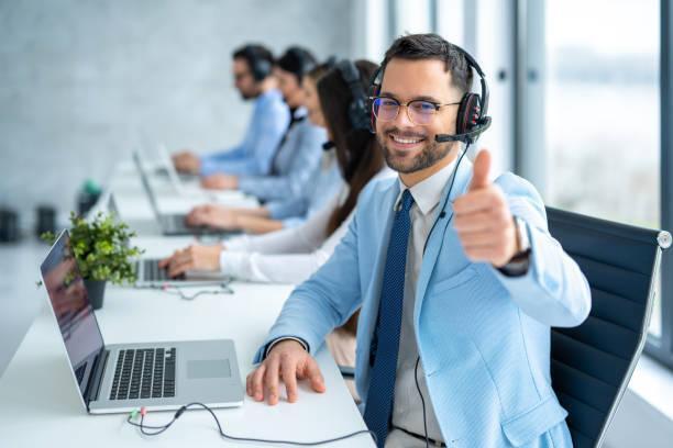 Male customer service agent showing approval sign with thumbs up while working in row with colleagues at call center Male customer service representative showing approval sign with thumbs up while working in a row with his other operators at call center office IT Support stock pictures, royalty-free photos & images