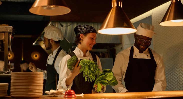 Chef in low lit kitchen holding fresh greens standing next to coworker prepping food